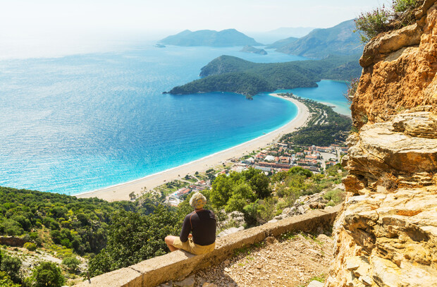 A person sits on a stone ledge overlooking a coastal landscape featuring a curved beach, turquoise sea, and distant green hills under a blue sky.